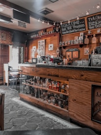 A cozy coffee shop interior featuring wooden decor, a counter with coffee brewing equipment, shelves stocked with jars and bottles, and signs displaying coffee-related messages. Ambient lighting with small string lights adds warmth to the atmosphere.
