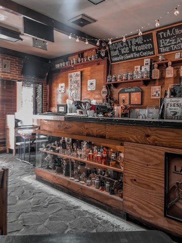 A cozy coffee shop interior featuring wooden decor, a counter with coffee brewing equipment, shelves stocked with jars and bottles, and signs displaying coffee-related messages. Ambient lighting with small string lights adds warmth to the atmosphere.