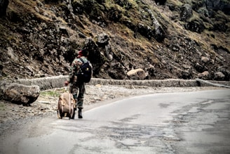 A person in camouflage attire walks along a winding mountain road carrying a large backpack and a duffle bag. The rocky terrain is covered with moss and sparse vegetation under a cloudy sky.