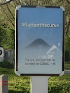 A public advertisement board with a message promoting social responsibility against COVID-19. It features the hashtag #flattenthecurve in bold white letters above a diagram illustrating a flattened curve. Below, the text 'Tous ensemble contre le COVID-19' is displayed, encouraging collective action. The board is set outdoors, surrounded by greenery.