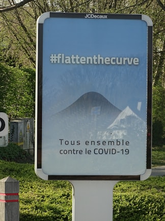 A public advertisement board with a message promoting social responsibility against COVID-19. It features the hashtag #flattenthecurve in bold white letters above a diagram illustrating a flattened curve. Below, the text 'Tous ensemble contre le COVID-19' is displayed, encouraging collective action. The board is set outdoors, surrounded by greenery.