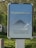 A public advertisement board with a message promoting social responsibility against COVID-19. It features the hashtag #flattenthecurve in bold white letters above a diagram illustrating a flattened curve. Below, the text 'Tous ensemble contre le COVID-19' is displayed, encouraging collective action. The board is set outdoors, surrounded by greenery.