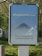 A public advertisement board with a message promoting social responsibility against COVID-19. It features the hashtag #flattenthecurve in bold white letters above a diagram illustrating a flattened curve. Below, the text 'Tous ensemble contre le COVID-19' is displayed, encouraging collective action. The board is set outdoors, surrounded by greenery.