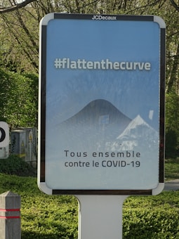A public advertisement board with a message promoting social responsibility against COVID-19. It features the hashtag #flattenthecurve in bold white letters above a diagram illustrating a flattened curve. Below, the text 'Tous ensemble contre le COVID-19' is displayed, encouraging collective action. The board is set outdoors, surrounded by greenery.