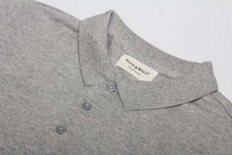 A neatly folded grey polo shirt with a collar and three buttons is displayed, featuring a label sewn inside the collar area with the brand name 'VITO & WILLY'. The fabric appears soft and finely woven, and the color is a consistent light grey.