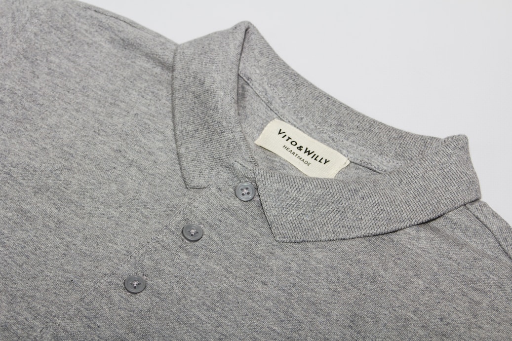 A neatly folded grey polo shirt with a collar and three buttons is displayed, featuring a label sewn inside the collar area with the brand name 'VITO & WILLY'. The fabric appears soft and finely woven, and the color is a consistent light grey.