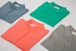 Four folded polo shirts in different colors are arranged symmetrically on a flat surface. The colors include blue, green, coral, and gray. Each shirt has a tag visible near the collar.