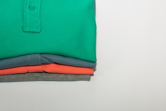 A stack of neatly folded polo shirts in four different colors, with the top shirt being green, followed by blue, coral, and grey. The shirts are arranged such that their collars and buttons are visible, indicating they are polo shirts. The background is a plain light color, highlighting the vividness of the folded shirts.