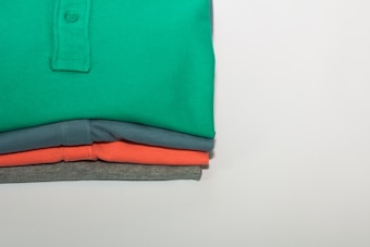 A stack of neatly folded polo shirts in four different colors, with the top shirt being green, followed by blue, coral, and grey. The shirts are arranged such that their collars and buttons are visible, indicating they are polo shirts. The background is a plain light color, highlighting the vividness of the folded shirts.