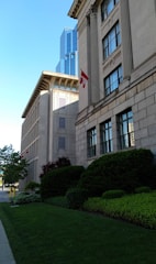 A city street with a prominent stone building featuring large windows and classical architectural details. In the foreground, well-manicured bushes and trees are visible alongside a neatly trimmed lawn. A modern glass skyscraper is seen in the background. The Canadian flag is displayed prominently on the stone building.