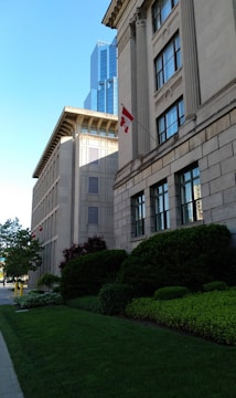 A city street with a prominent stone building featuring large windows and classical architectural details. In the foreground, well-manicured bushes and trees are visible alongside a neatly trimmed lawn. A modern glass skyscraper is seen in the background. The Canadian flag is displayed prominently on the stone building.