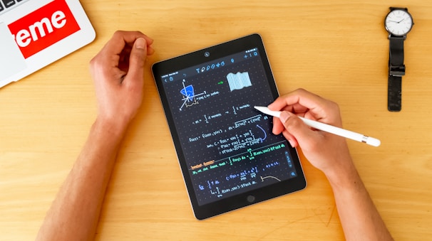 A person uses a stylus to write mathematical equations on a tablet, which is placed on a wooden table. The tablet screen displays complex graphs and formulas. Nearby, there is a wristwatch and a laptop with a prominent 'eme' sticker. The person's left hand is resting on the table while the right hand holds the stylus.
