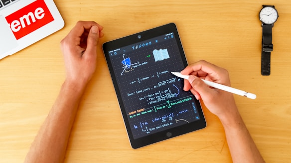 A person uses a stylus to write mathematical equations on a tablet, which is placed on a wooden table. The tablet screen displays complex graphs and formulas. Nearby, there is a wristwatch and a laptop with a prominent 'eme' sticker. The person's left hand is resting on the table while the right hand holds the stylus.