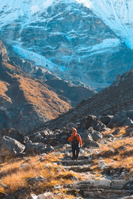 A hiker in orange and black outdoor gear is trekking along a rocky path amidst mountainous terrain. The background is dominated by a towering, snow-covered mountain range, contrasting with the rugged, grassy hills in the foreground. Sunlight creates a striking play of light and shadow across the landscape.