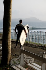 A surfer in a wetsuit stands on a cobbled pathway near the ocean, holding a surfboard and looking towards the waves. A white, ornate fence separates the path from the sea, and benches are situated along the pathway. A tree trunk on the left adds a natural element to the scene while the distant horizon is slightly hazy with hills in the background.