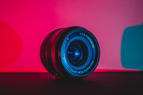 A camera lens is positioned on a surface, illuminated by vibrant red and teal lighting, creating a dramatic and modern aesthetic. The lens, marked with specifications, is the focal point against a contrasting background.