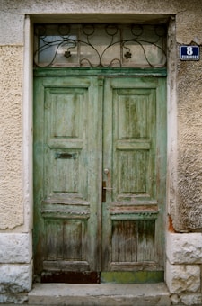 An old, weathered double wooden door with a green patina, set into a stone building. The door has intricate wrought iron details on a small window above it, and a small metal mail slot is visible. The stone around the door has a rough texture, and a wall plaque displays the number '8' along with the word 'PLINARSKA'.