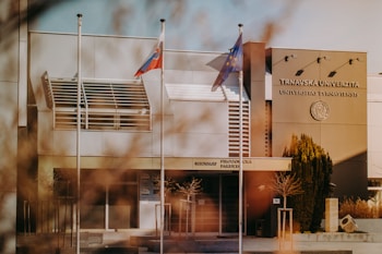 A modern university building with three flagpoles, two displaying flags, in front of its entrance. The building has a sleek design with clean lines and a neutral color palette. The name 'Trnavska Univerzita' and a seal are visible on its facade.