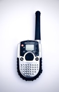 A handheld radio device with a black antenna, silver body, and numerous small buttons, including a push-to-talk button and a menu button. The screen is small and above the buttons, with the brand name visible below.