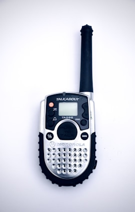 A handheld radio device with a black antenna, silver body, and numerous small buttons, including a push-to-talk button and a menu button. The screen is small and above the buttons, with the brand name visible below.