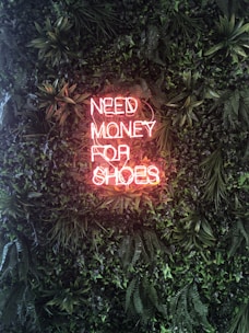 A neon sign displaying the words 'NEED MONEY FOR SHOES' is prominently set against a lush green wall covered with abundant foliage. The neon sign emits a warm, pinkish glow that contrasts with the rich, vibrant greens of the plants.