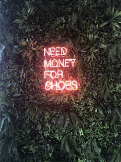 A neon sign displaying the words 'NEED MONEY FOR SHOES' is prominently set against a lush green wall covered with abundant foliage. The neon sign emits a warm, pinkish glow that contrasts with the rich, vibrant greens of the plants.