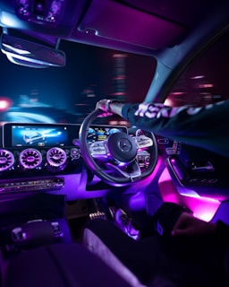 A high-tech car interior featuring a sleek dashboard with digital displays, vibrant neon lighting, and a modern steering wheel. The ambient lighting casts a purple and blue glow throughout the cabin, creating a futuristic atmosphere. A person is driving the car, and the cityscape outside appears blurred, indicating motion.