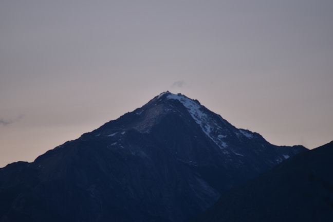 A mountain peak silhouetted against a twilight sky, with a faint figure barely visible near the summit.