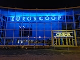 A modern building facade with a large glass frontage illuminated by bright blue and yellow neon lights. The word 'Euroscoop' is displayed at the top, while the entrance is marked by the word 'Cinema'. Bicycles are seen parked in front of the building, and the overall atmosphere is urban and contemporary.
