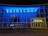 A modern building facade with a large glass frontage illuminated by bright blue and yellow neon lights. The word 'Euroscoop' is displayed at the top, while the entrance is marked by the word 'Cinema'. Bicycles are seen parked in front of the building, and the overall atmosphere is urban and contemporary.