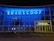 A modern building facade with a large glass frontage illuminated by bright blue and yellow neon lights. The word 'Euroscoop' is displayed at the top, while the entrance is marked by the word 'Cinema'. Bicycles are seen parked in front of the building, and the overall atmosphere is urban and contemporary.