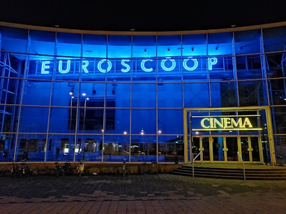A modern building facade with a large glass frontage illuminated by bright blue and yellow neon lights. The word 'Euroscoop' is displayed at the top, while the entrance is marked by the word 'Cinema'. Bicycles are seen parked in front of the building, and the overall atmosphere is urban and contemporary.