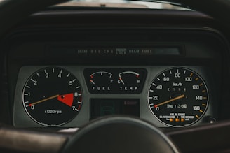 A detailed view of a car dashboard displaying various gauges, including a tachometer, fuel gauge, temperature gauge, and speedometer. The tachometer indicates engine speed in revolutions per minute, with a highlighted red zone. The fuel gauge shows fuel levels, and the temperature gauge reads engine temperature. The speedometer measures speed in kilometers per hour, showing vehicle mileage.