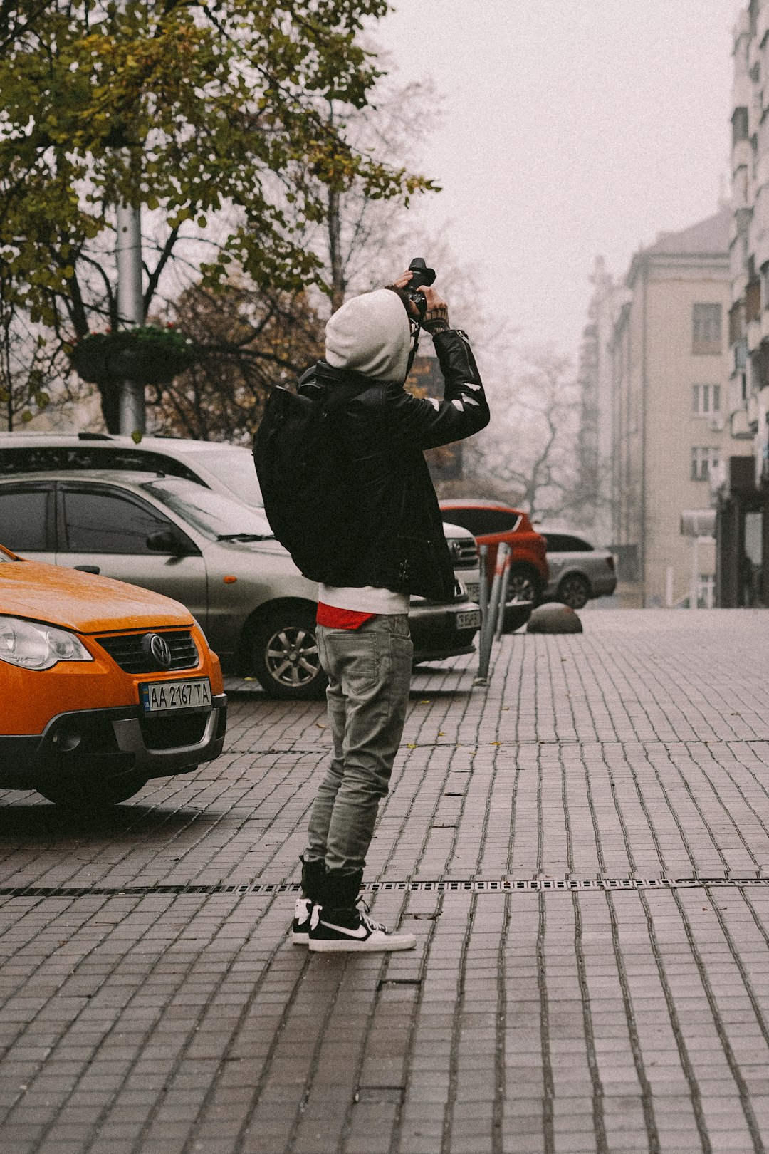 man in black jacket and gray pants taking photo of orange car during daytime