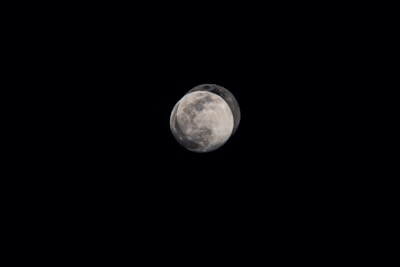 A celestial object resembling the moon appears against a pitch-black sky. The image captures two overlapping views of the moon, suggesting a time-lapse or motion effect. The surface shows distinct craters and textures typical of the lunar surface.