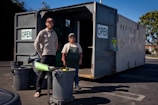 Two people standing in front of an open recycling center container with multiple trash bins nearby. The container has signs indicating that it is open for business. The scene is set outdoors in a parking lot area, under clear blue skies.