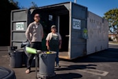 Two people standing in front of an open recycling center container with multiple trash bins nearby. The container has signs indicating that it is open for business. The scene is set outdoors in a parking lot area, under clear blue skies.