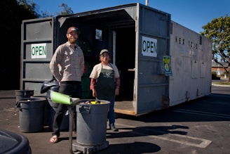 Two people standing in front of an open recycling center container with multiple trash bins nearby. The container has signs indicating that it is open for business. The scene is set outdoors in a parking lot area, under clear blue skies.