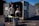 Two people standing in front of an open recycling center container with multiple trash bins nearby. The container has signs indicating that it is open for business. The scene is set outdoors in a parking lot area, under clear blue skies.