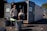 Two people standing in front of an open recycling center container with multiple trash bins nearby. The container has signs indicating that it is open for business. The scene is set outdoors in a parking lot area, under clear blue skies.