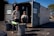 Two people standing in front of an open recycling center container with multiple trash bins nearby. The container has signs indicating that it is open for business. The scene is set outdoors in a parking lot area, under clear blue skies.