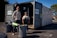 Two people standing in front of an open recycling center container with multiple trash bins nearby. The container has signs indicating that it is open for business. The scene is set outdoors in a parking lot area, under clear blue skies.