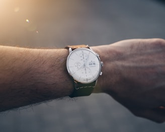A close-up of a wrist with a luxury analog watch featuring a white face and a tan leather strap. The watch shows various small dials and markers, indicating detailed timing functions. The wrist is slightly hairy, and the background appears to be blurred, with soft sunlight illuminating part of the image.