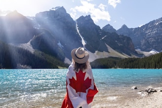 A person wearing a hat draped in a Canadian flag stands on the shore of a clear, turquoise lake surrounded by rugged, snow-capped mountains. Sunlight streams through partially cloudy skies, casting light on the mountains and lake.