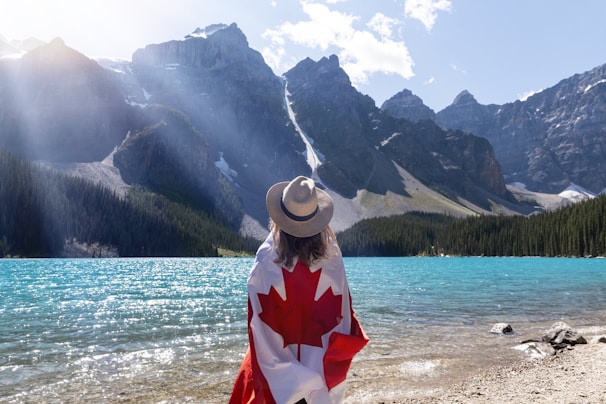 A person wearing a hat draped in a Canadian flag stands on the shore of a clear, turquoise lake surrounded by rugged, snow-capped mountains. Sunlight streams through partially cloudy skies, casting light on the mountains and lake.