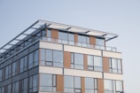 A modern building with large glass windows and horizontal wooden panels. The structure features a rooftop area with a glass railing and a minimalistic design. The architecture is contemporary with clean lines and a neutral color scheme.