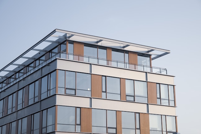 A modern building with large glass windows and horizontal wooden panels. The structure features a rooftop area with a glass railing and a minimalistic design. The architecture is contemporary with clean lines and a neutral color scheme.