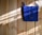 A bright blue crocheted bag with two wooden buttons is hanging against a light wooden wall. Sunlight filters in, casting shadows and highlighting the texture of the crochet work.