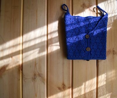 A bright blue crocheted bag with two wooden buttons is hanging against a light wooden wall. Sunlight filters in, casting shadows and highlighting the texture of the crochet work.