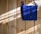 A bright blue crocheted bag with two wooden buttons is hanging against a light wooden wall. Sunlight filters in, casting shadows and highlighting the texture of the crochet work.